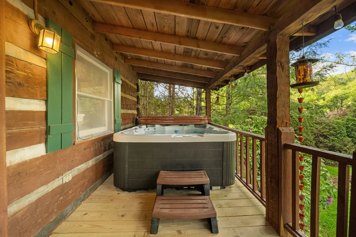 A tranquil deck scene featuring a bubbling hot tub set against the wooden exterior of the cabin, with green shutters and a hanging lantern lending rustic charm. The deck opens to the outdoors, suggesting space for morning coffee or stargazing soaks.