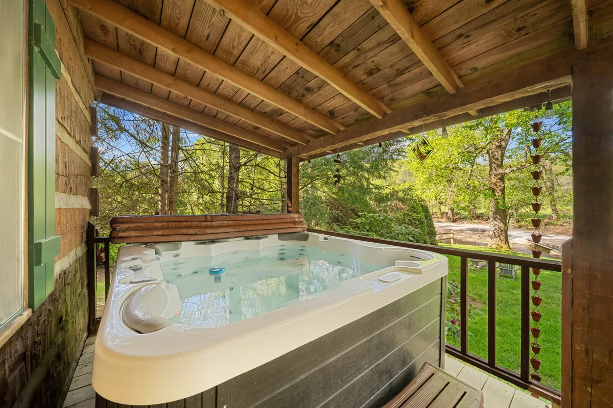 The image showcases a serene exterior scene featuring a steaming hot tub set on a wooden deck, surrounded by tall trees. The peaceful, natural environment is ideal for relaxing after a day of adventure, with views for stargazing or enjoying a sunrise coffee.
