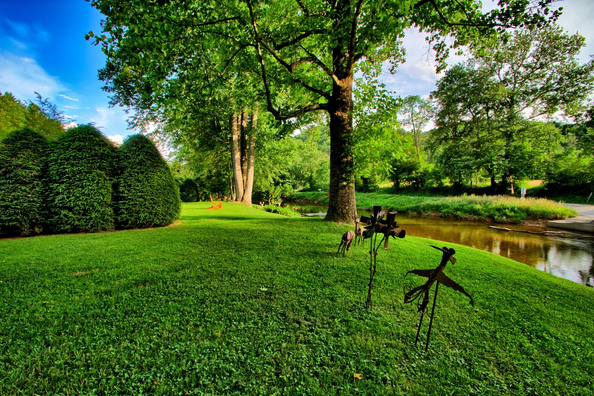 A grassy yard bordered by large green trees and a calm stream, with whimsical sculptures dotting the lawn. The peaceful setting invites strolling, picnicking, and quiet play by the water’s edge.