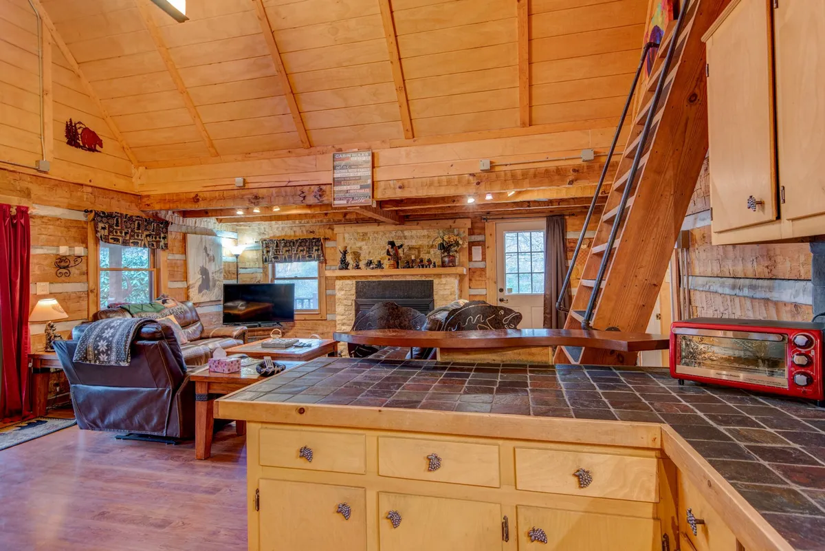 A warm, open-plan living area featuring a cozy fireplace flanked by wooden beams, a spacious sectional sofa, a rustic kitchen counter, and a ladder leading to a loft. Large windows allow natural light to flood the room, subtly connecting the interior with the outside scenery.