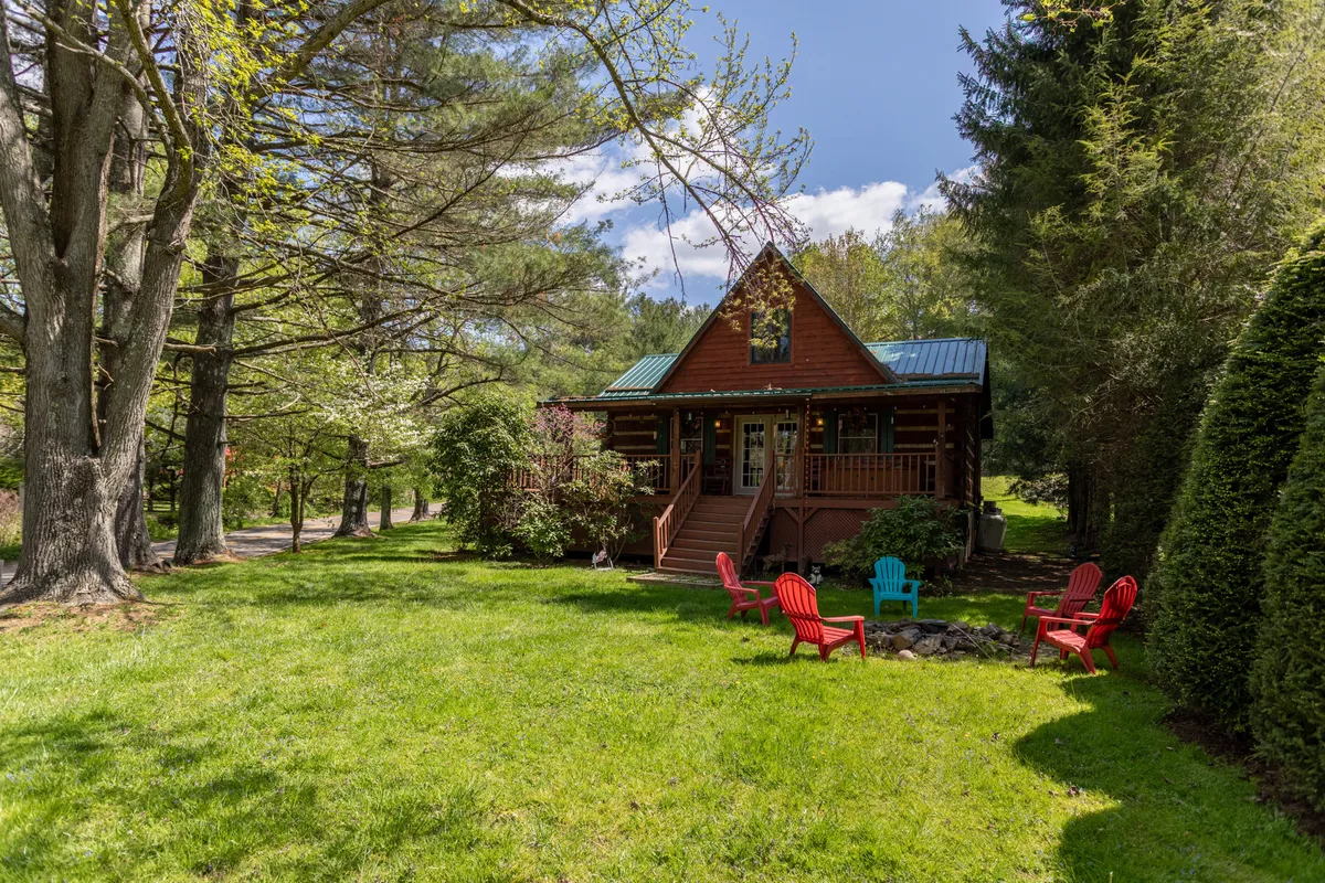 This image features a log cabin set beside a lush, flat green lawn, accented by red Adirondack chairs and a fire pit. The expansive yard provides ample open space and is bordered by trees, creating a welcoming and safe play area for both kids and dogs. The fire pit area, with its seating, suggests opportunities for evening gatherings and making s’mores.