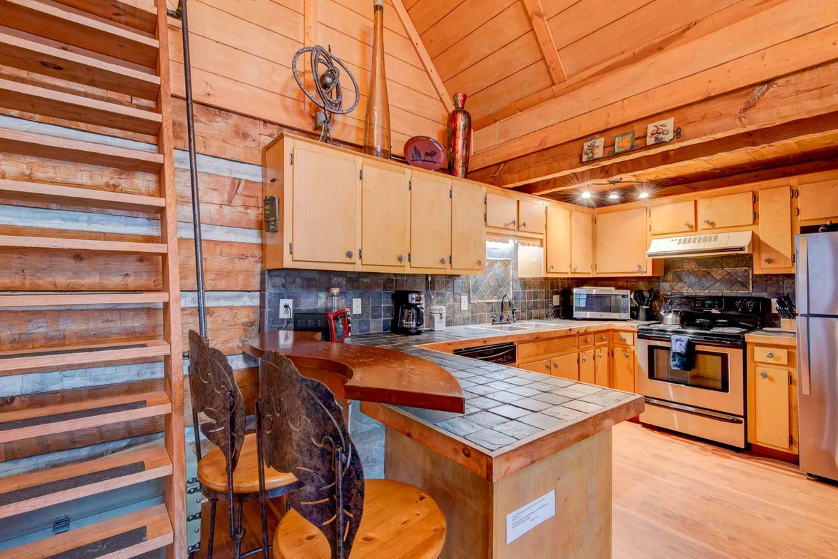 This image features a kitchen area with a kitchen island and bar stools, visible cabinets and appliances, and a wooden staircase in the background. The setup is open and inviting, ideal for group cooking or casual gatherings. The combination of modern appliances and rustic touches highlights the kitchen's role as a central, functional, and social space.