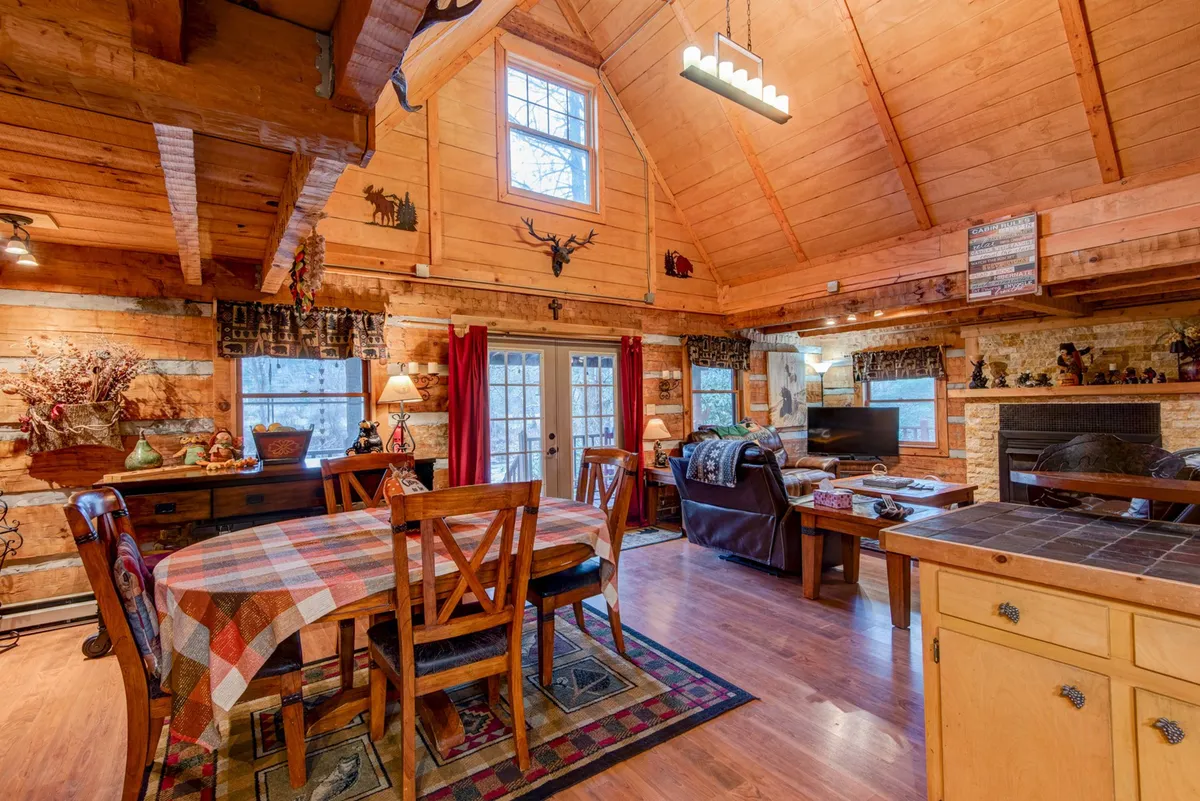 A welcoming dining space with a checkered tablecloth, ample natural light streaming through large windows, and comfortable seating. The scene is accented by rustic decorative elements, reinforcing the cabin’s mountain character and the flow between interior and exterior worlds.