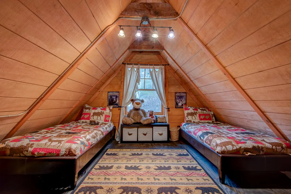 A playful loft area featuring two twin beds, a patterned rug, a large teddy bear, and a window with curtains. The space feels comfortable, fun, and welcoming, making it perfect for children or solo travelers seeking a cozy hideaway.