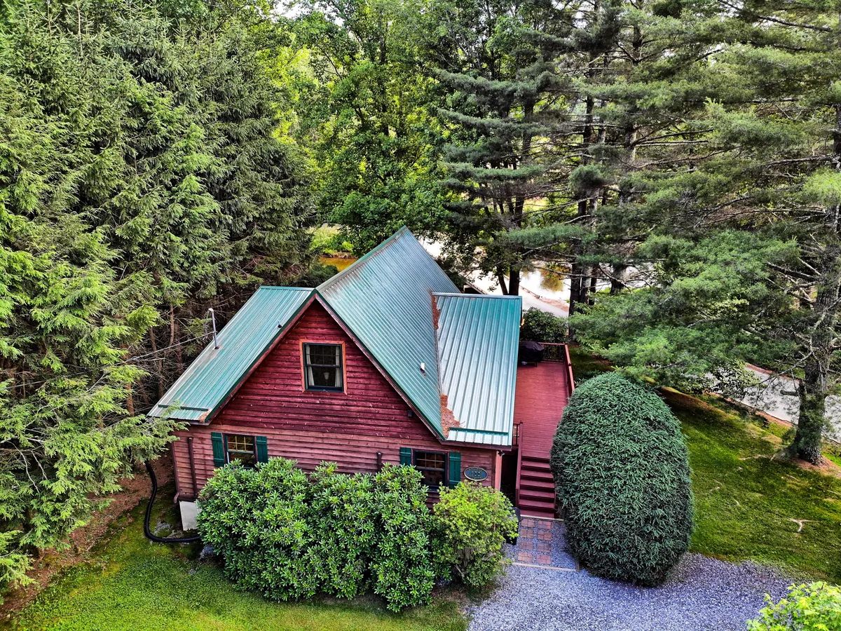 This exterior shot features the rustic red cabin with a green metal roof, nestled among lush trees. The image captures the peaceful setting with a pathway leading toward the entrance, inviting guests to explore the surrounding nature or relax on the porch.