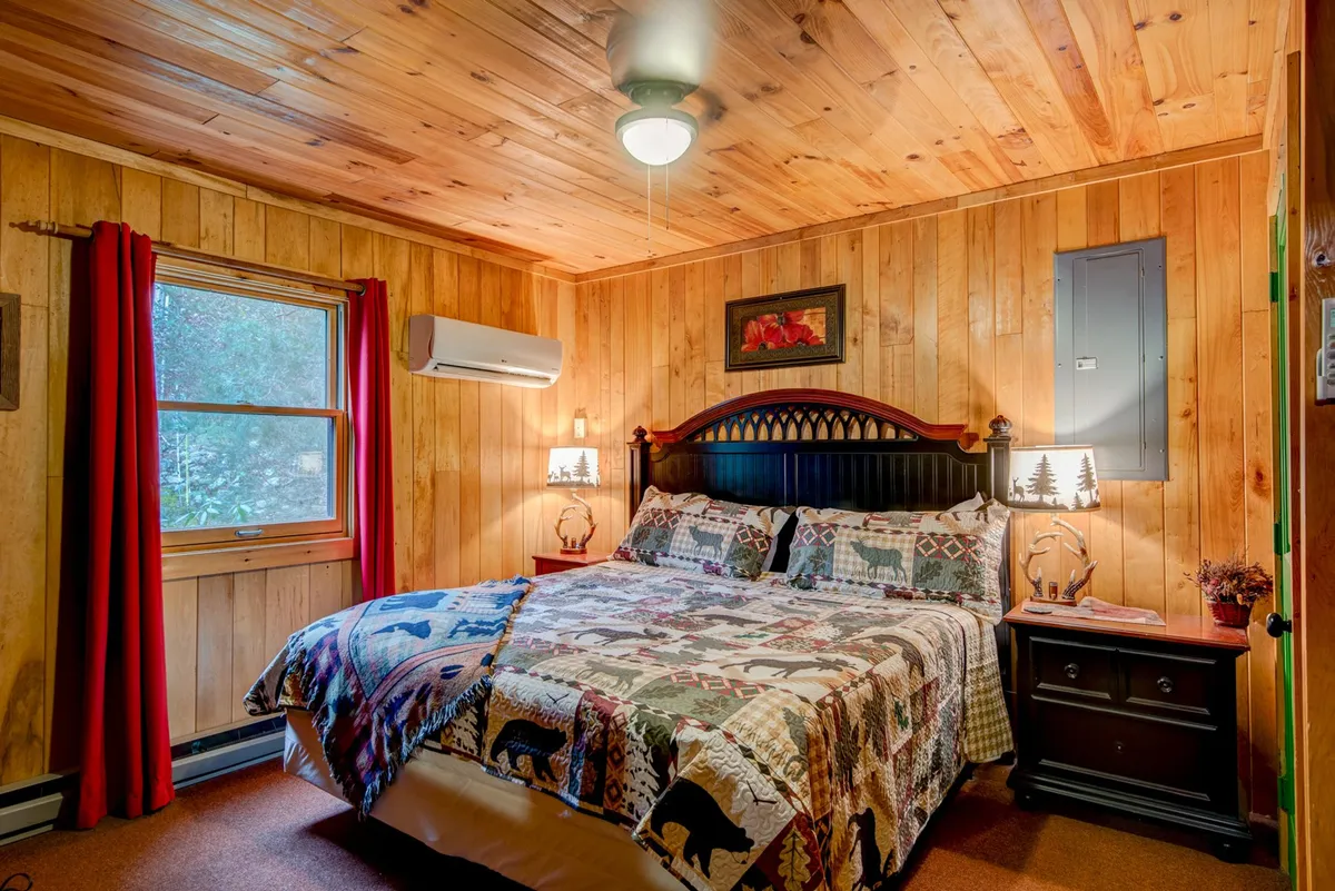 A warmly lit bedroom with a wooden-paneled wall, a bed dressed in a patterned quilt, nightstands with lamps on either side, and a window adorned with red curtains letting in the morning sunlight. The overall atmosphere is inviting and restful, emphasizing natural textures and soft lighting.