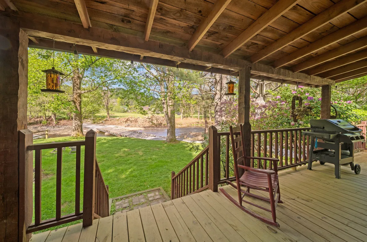 A wooden deck overlooking the gently flowing stream, featuring a rocking chair, grill, and surrounded by lush greenery. The setting is tranquil, with ample space for outdoor relaxation or family gatherings right at the water’s edge.
