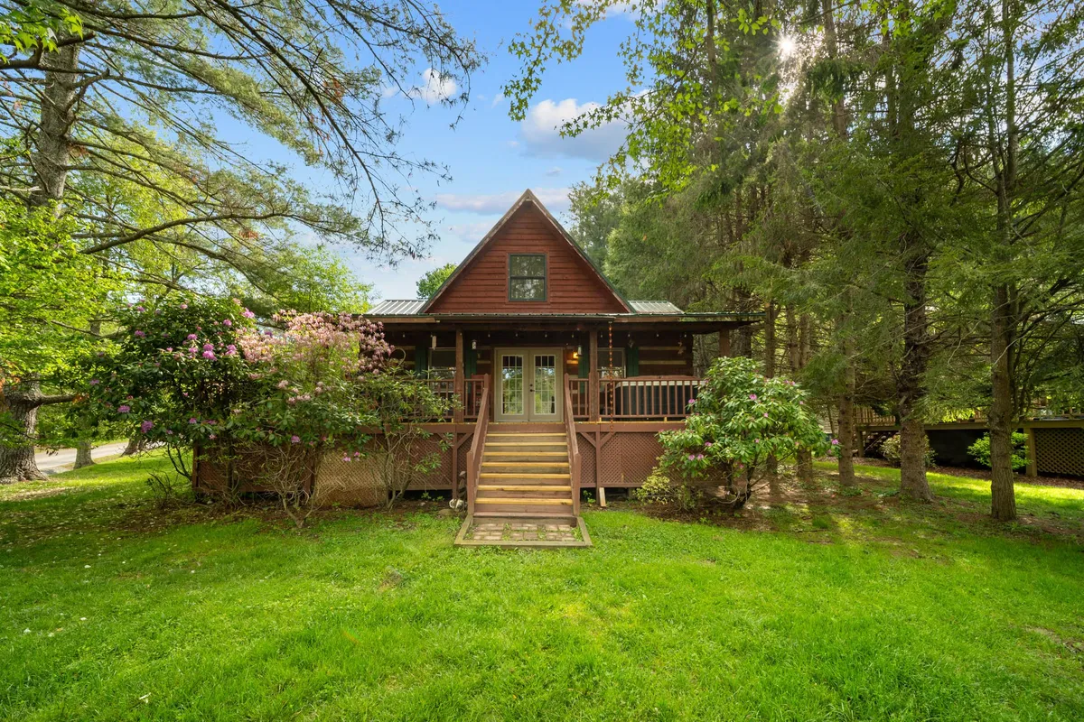 A welcoming front porch with wooden stairs, surrounded by vibrant greenery and flowering bushes. The porch features comfortable seating, inviting families to relax while enjoying the view of the yard and the peaceful setting. The natural landscaping frames the cabin and hints at the play spaces beyond.