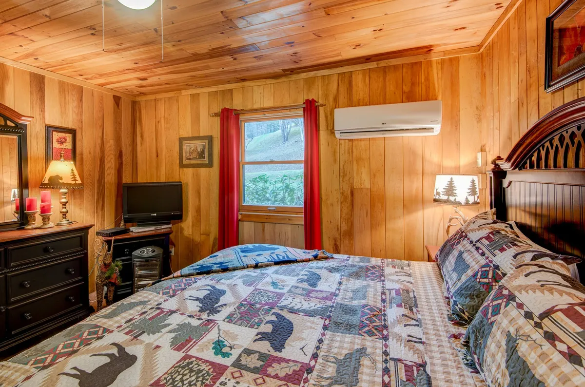 A welcoming bedroom with a patterned quilt on the bed, a window framed by red curtains, and a dresser with a mirror and lamp. The setting suggests a comfortable space ideal for relaxing, reading, or enjoying a quiet moment.