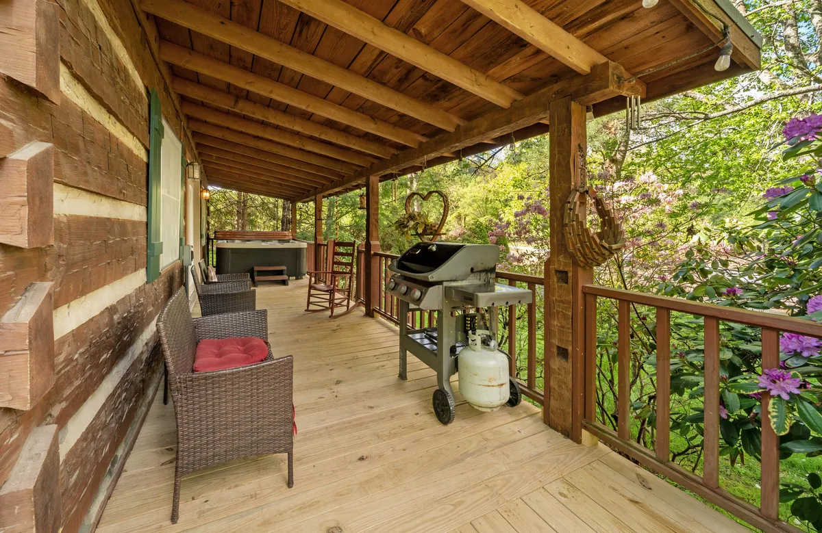 The welcoming exterior of the cabin with wooden log walls, a grill, rocking chairs, and vibrant potted flowers. The setting radiates warmth and invites visitors to relax outdoors, surrounded by nature.
